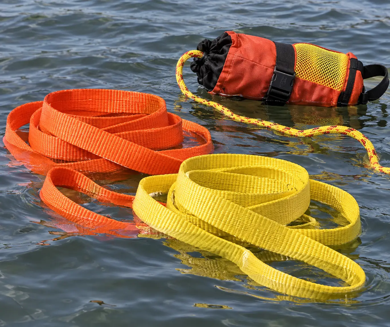 High-visibility orange and yellow floating polypropylene webbing straps coiled on the water surface next to a throw bag rescue line, demonstrating buoyant webbing used in water rescue equipment.